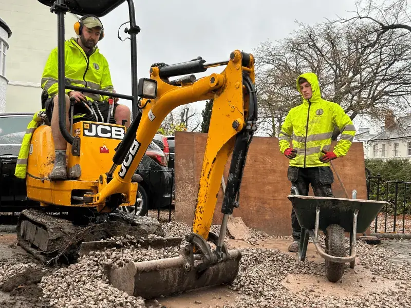 Two Silver Tree Services operatives; one is sat on a JCB mini-digger and the other is stood behind a wheelbarrow next to the JCB.